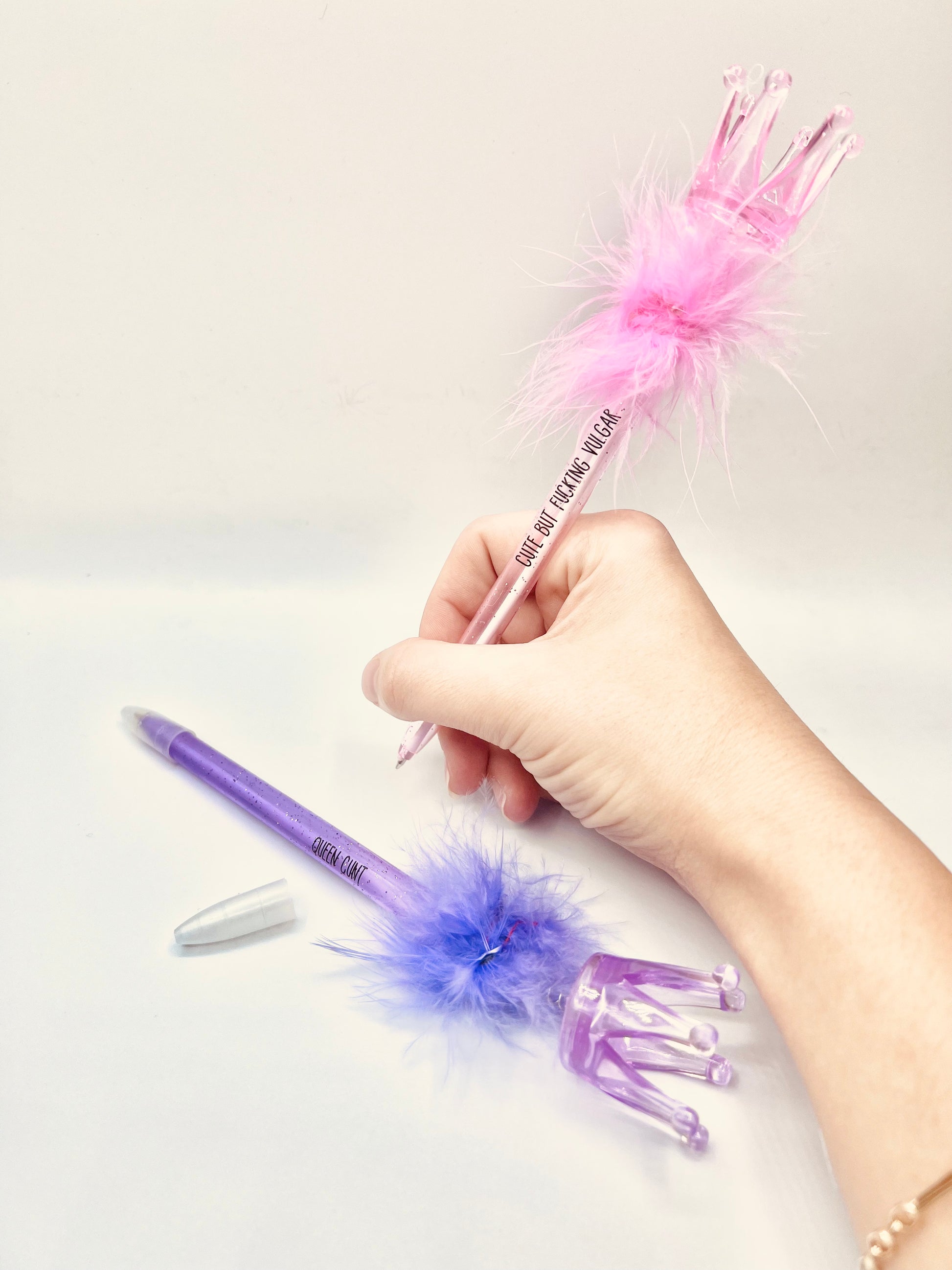 2 pack pens, pink and purple. Pink pen says 'cute but fucking vulgar' the purple says 'queen cunt'. Both pens have a glittery base, fluffy top and a plastic crown topper. Blue ink