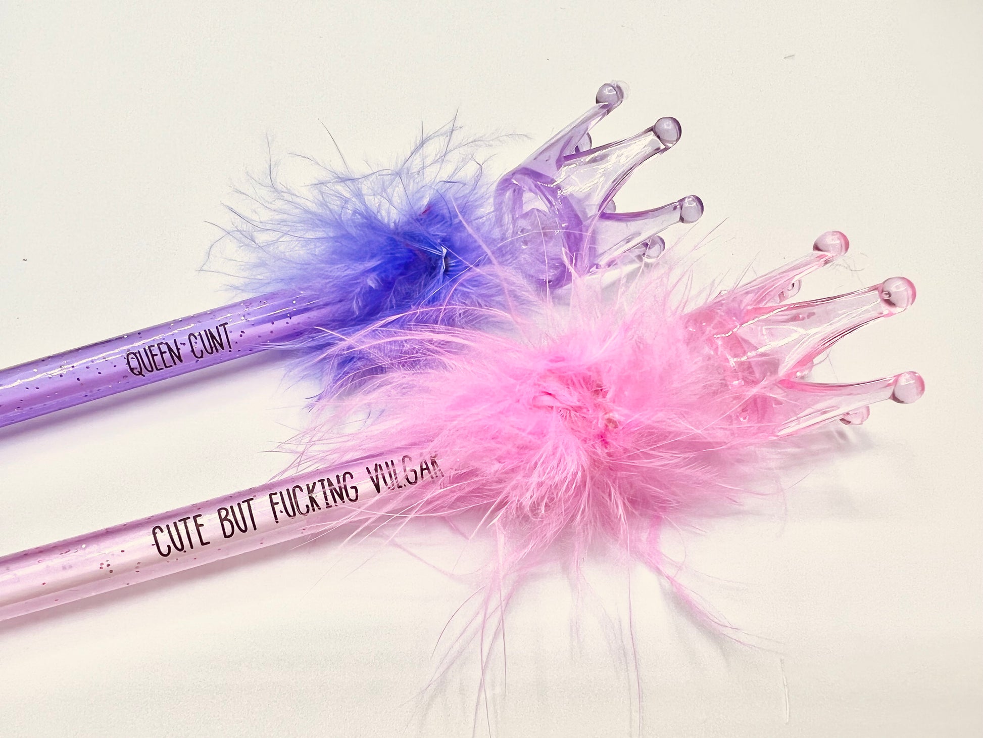 2 pack pens, pink and purple. Pink pen says 'cute but fucking vulgar' the purple says 'queen cunt'. Both pens have a glittery base, fluffy top and a plastic crown topper. Blue ink