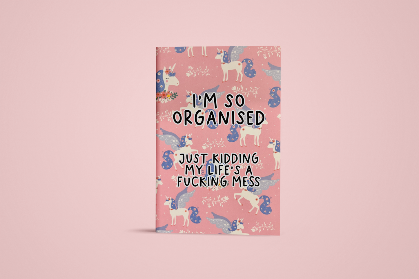 Pink notebook with a continuous unicorn design and in the middle it reads 'i'm so organised, just kidding my life's a fucking mess' in black ink.