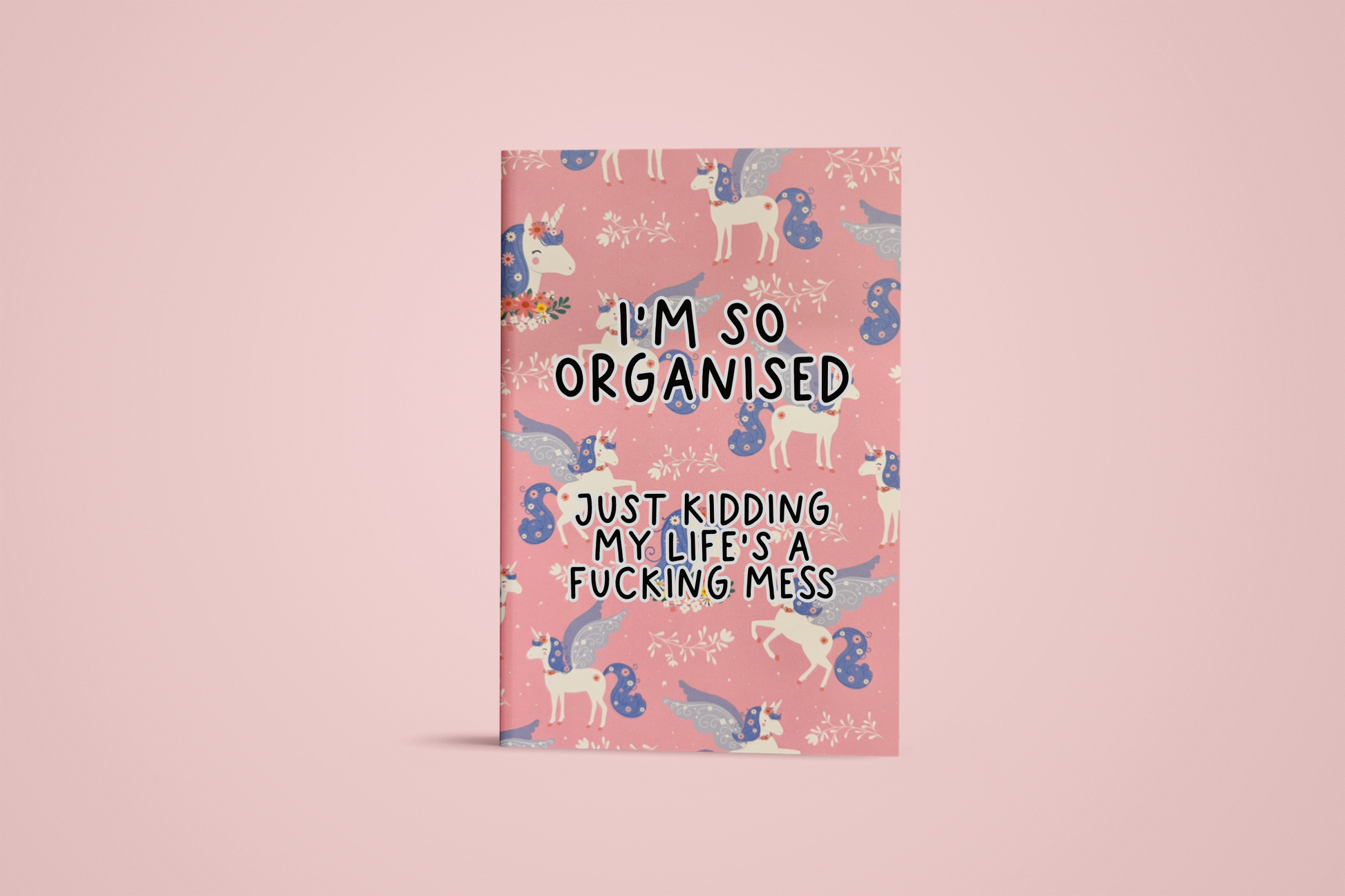 Pink notebook with a continuous unicorn design and in the middle it reads 'i'm so organised, just kidding my life's a fucking mess' in black ink.