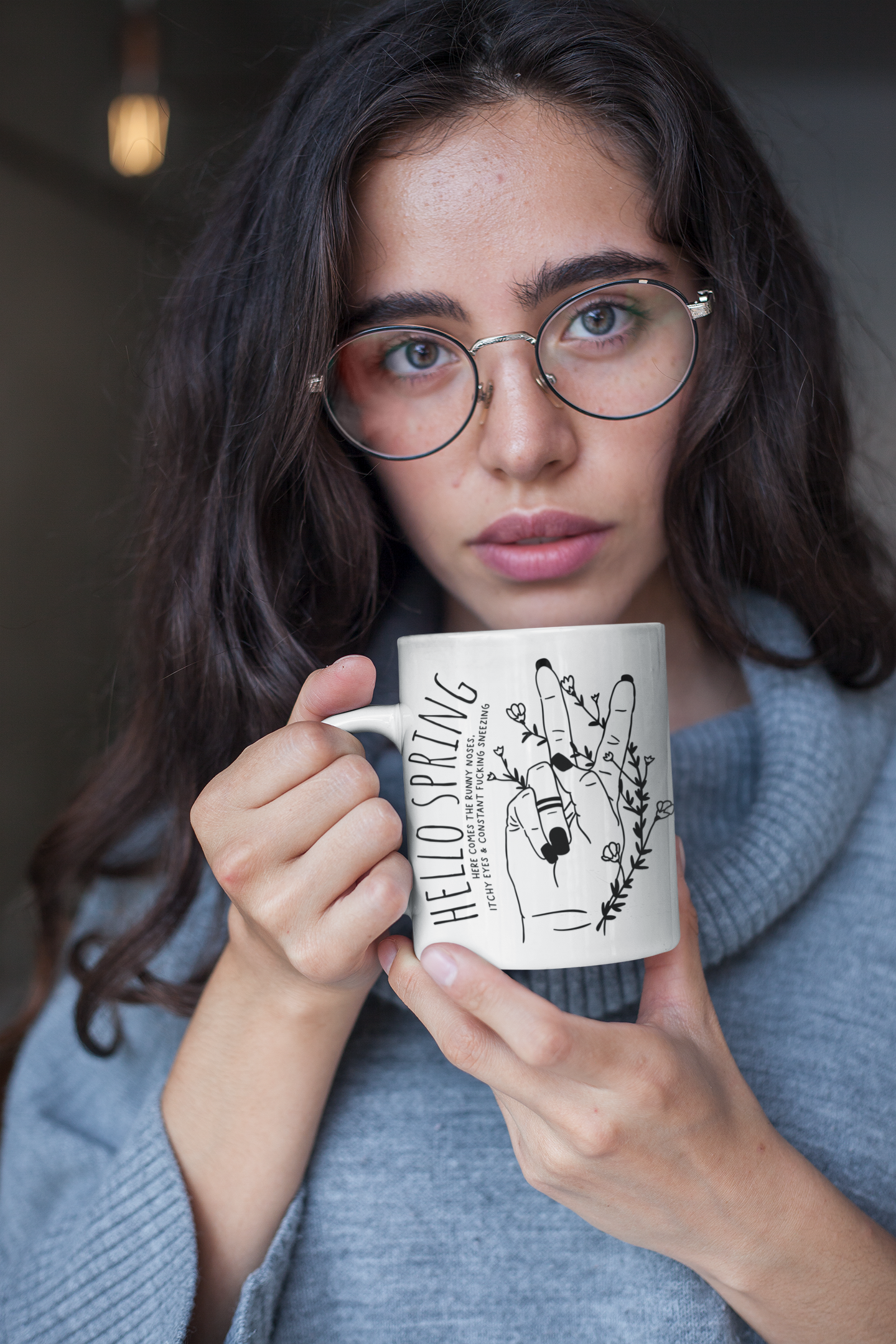 White ceramic mug with a cute boho style drawing of a hand sticking two fingers up, covered in twining flowers. There is a quote to the left which reads ' hello spring - here comes the runny noses, itchy eyes & constant fucking sneezing' printed in black.