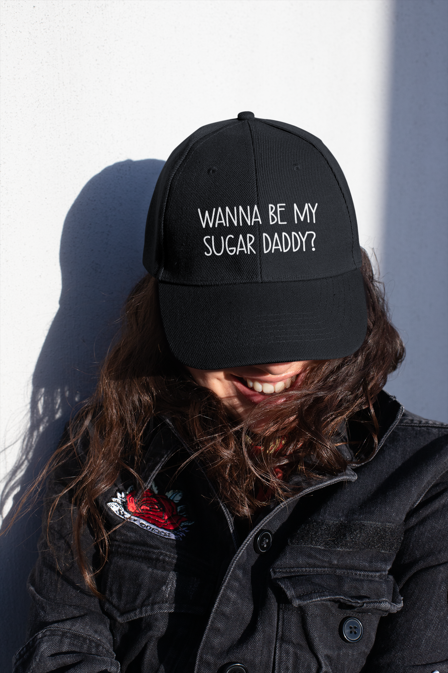 A lady wearing a black cotton cap with white lettering to the front which reads 'wanna be my sugar daddy?'.