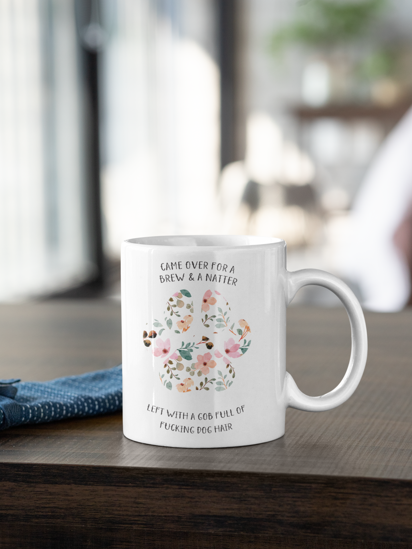White ceramic mug with a cute colourful floral dog paw print design. To the top of the print it reads 'came over for a brew & a natter' then to the bottom is says 'left with a gob full of fucking dog hair'. Printed in black ink.