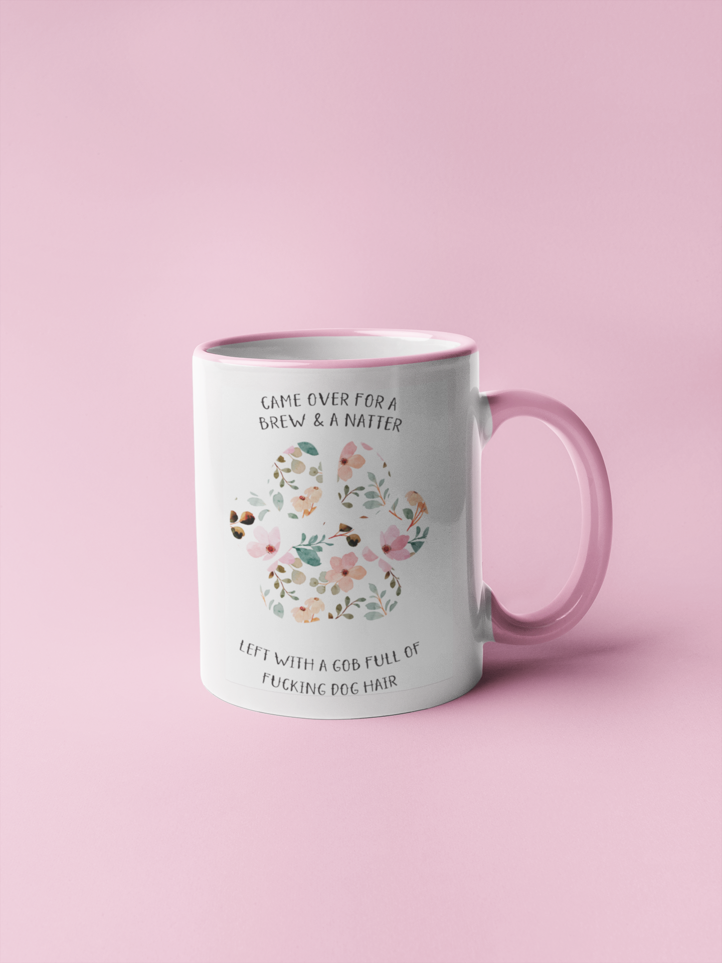 White mug with a pink handle and rim with a cute colourful floral dog paw print design. To the top of the print it reads 'came over for a brew & a natter' then to the bottom is says 'left with a gob full of fucking dog hair'. Printed in black ink.