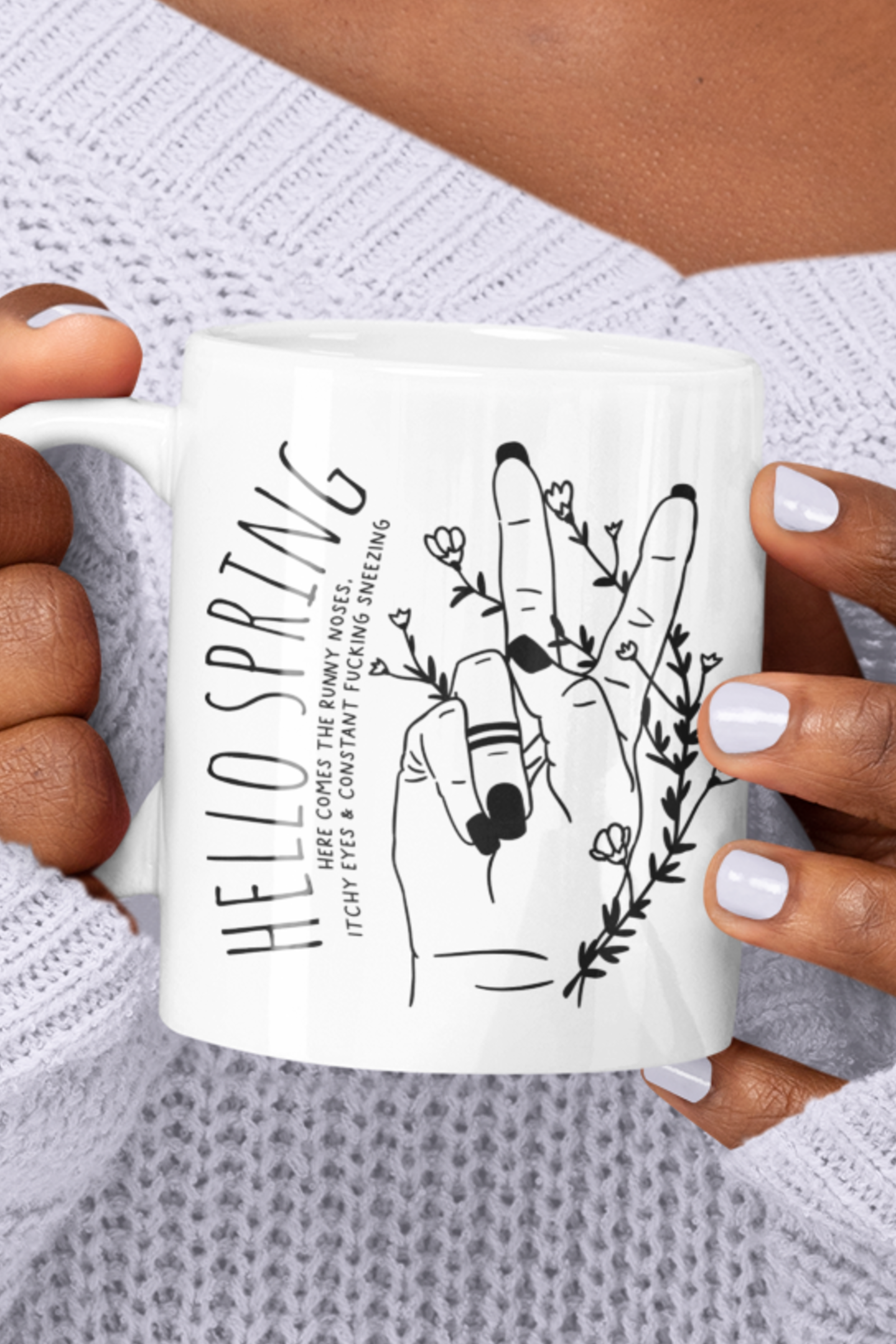 White ceramic mug with a cute boho style drawing of a hand sticking two fingers up, covered in twining flowers. There is a quote to the left which reads ' hello spring - here comes the runny noses, itchy eyes & constant fucking sneezing' printed in black.