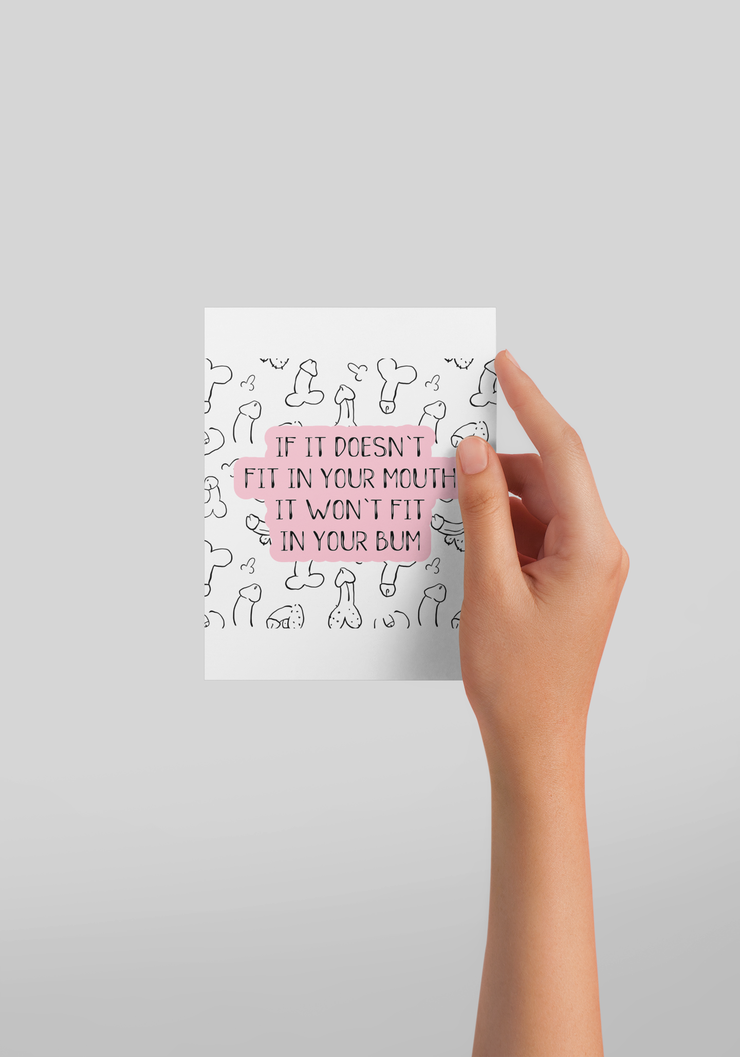 Girls hand holding a white vertical greetings card featuring a fun willy design print to the front. Over the print is a funny quote 'if it won't fit in your mouth, it won't fit in your bum', printed in black & pink ink