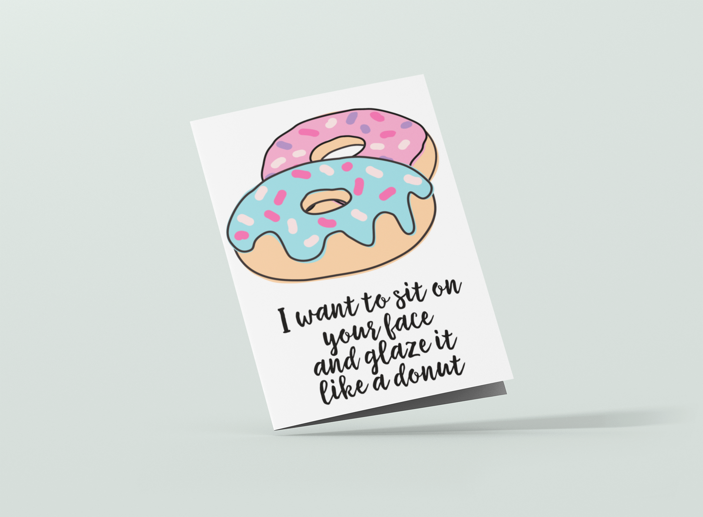 White vertical greetings card featuring a colourful donut design. To the bottom it reads 'i want to sit on your face & glaze it like a donut'. Printed in black ink.