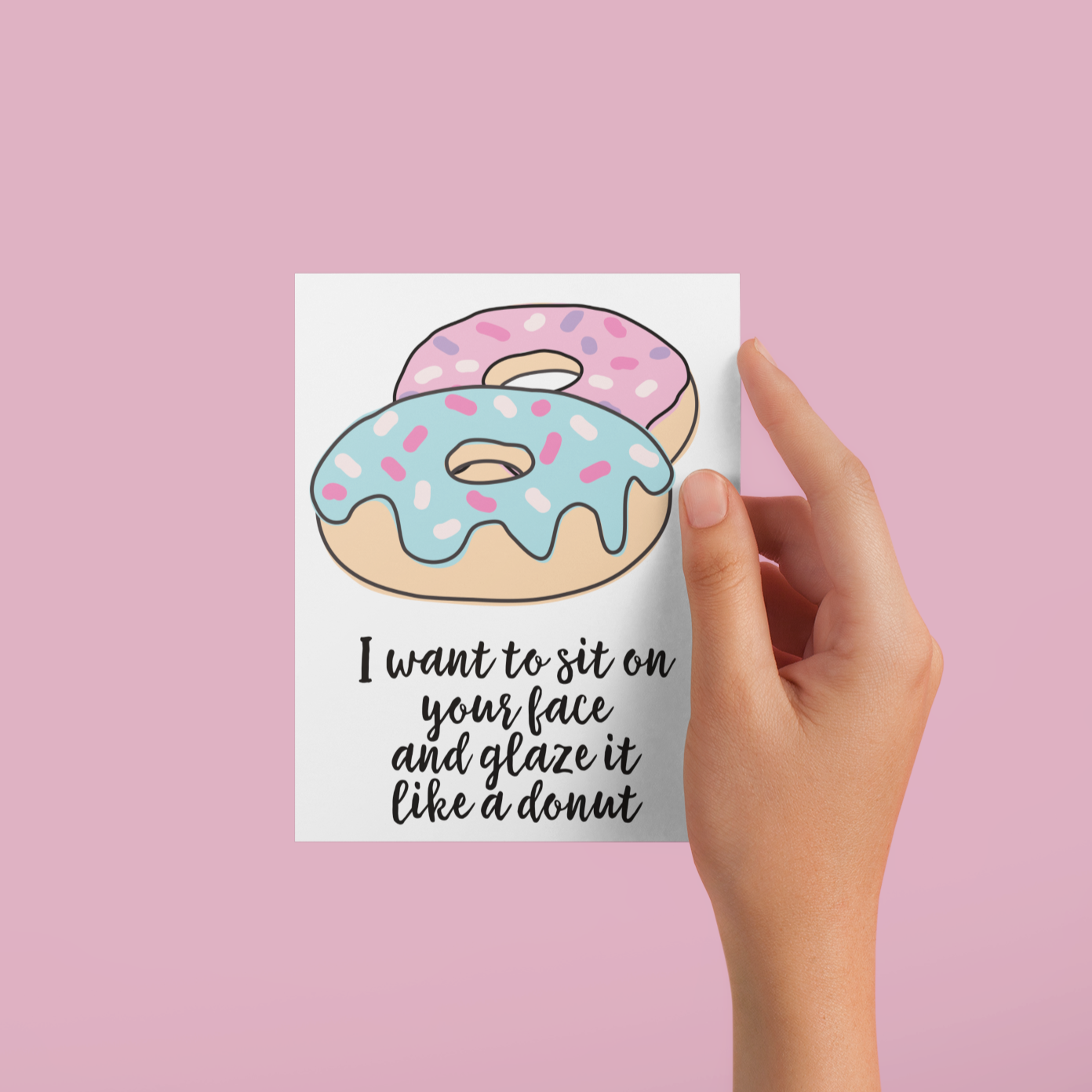 Hand holding a vertical greetings card featuring a colourful donut design. To the bottom it reads 'i want to sit on your face & glaze it like a donut'. Printed in black ink.