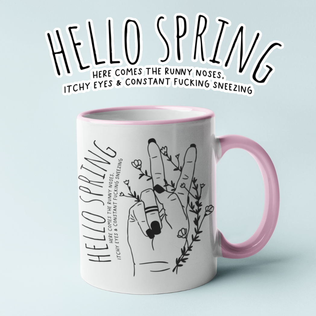 White ceramic mug with a pink handle and a cute boho style drawing of a hand sticking two fingers up, covered in twining flowers. There is a quote to the left which reads ' hello spring - here comes the runny noses, itchy eyes & constant fucking sneezing' printed in black.