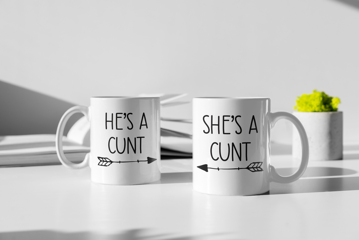 A pair of white ceramic mugs. One has 'he's a c*nt' with an arrow underneath pointing to the right. The other has 'she's a c*nt' with the arrow pointing to the left. Both printed to the front in black ink.