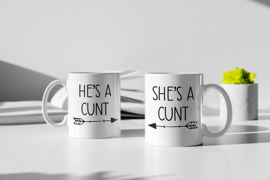 A pair of white ceramic mugs. One has 'he's a c*nt' with an arrow underneath pointing to the right. The other has 'she's a c*nt' with the arrow pointing to the left. Both printed to the front in black ink.