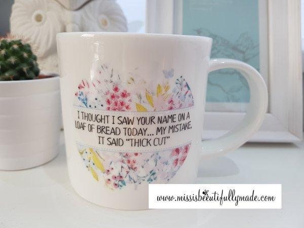 A white ceramic mug featuring the funny quote i thought i saw your name on a loaf of bread today My mistake, it said thick cut. Printed in black ink surrounded by a colourful floral design.