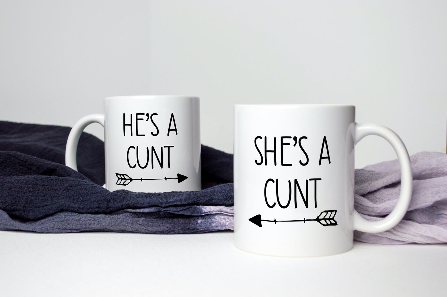 A pair of white ceramic mugs. One has 'he's a c*nt' with an arrow underneath pointing to the right. The other has 'she's a c*nt' with the arrow pointing to the left. Both printed to the front in black ink.