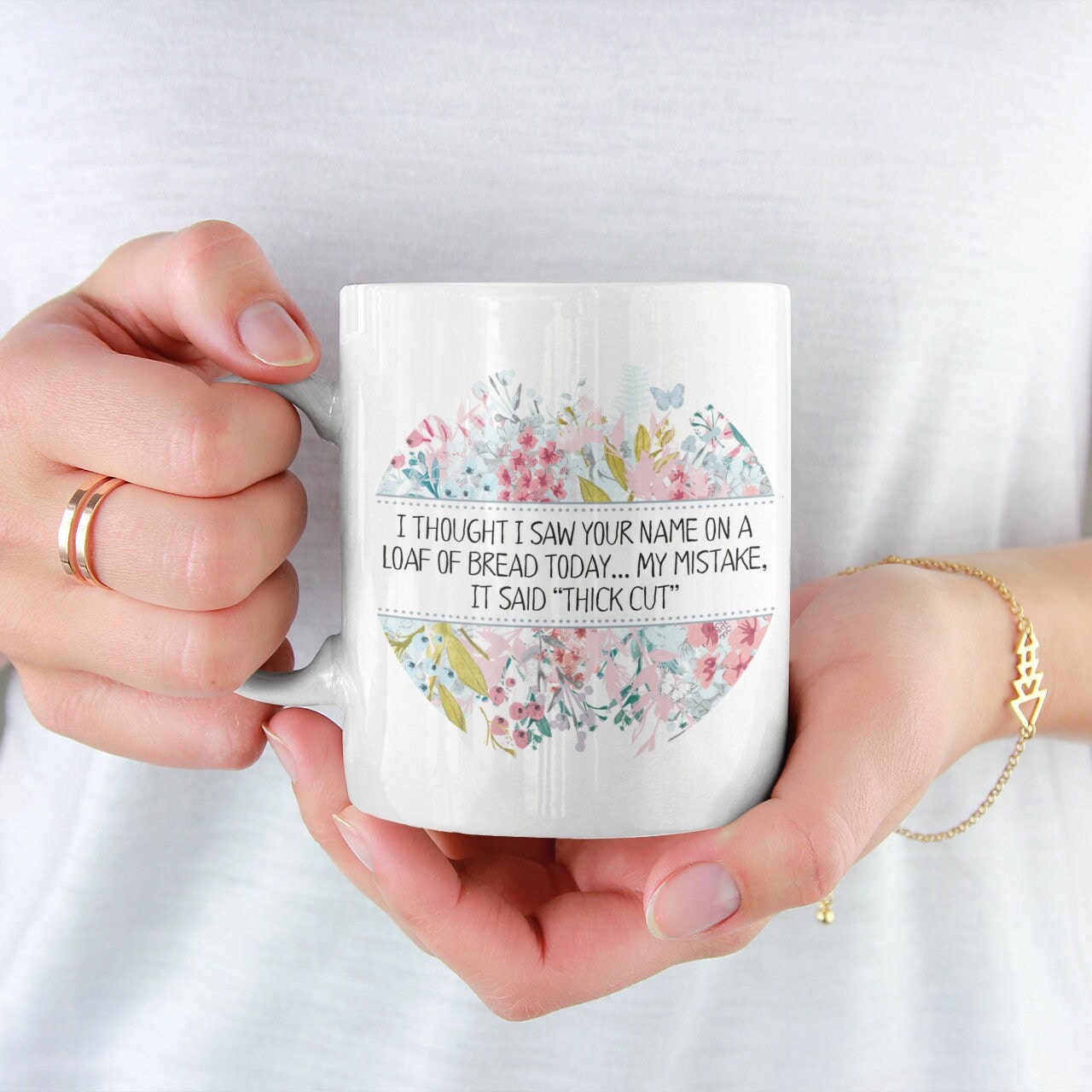 A white ceramic mug featuring the funny quote i thought i saw your name on a loaf of bread today My mistake, it said thick cut. Printed in black ink surrounded by a colourful floral design.
