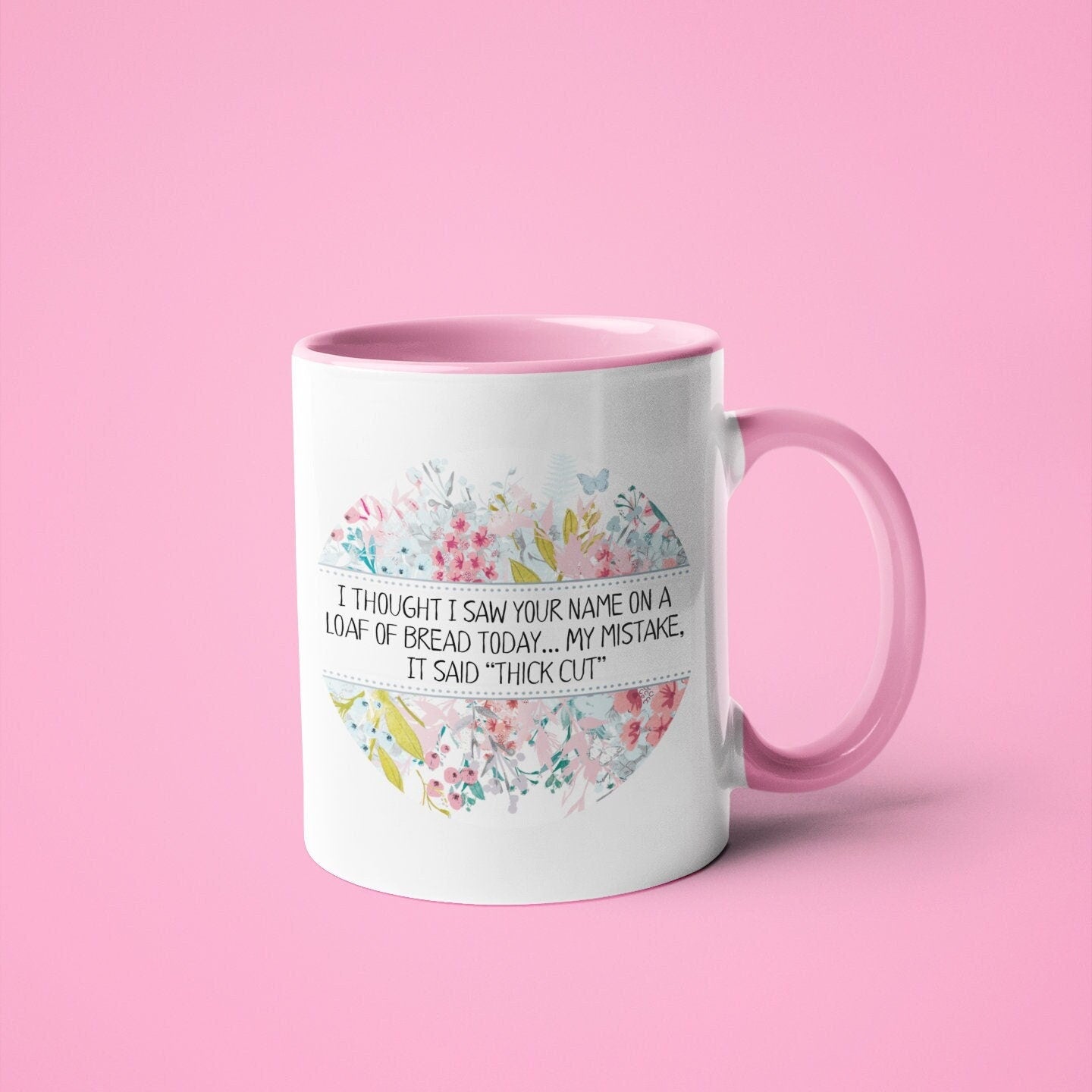A white ceramic mug with a pink handle & inner featuring the funny quote i thought i saw your name on a loaf of bread today My mistake, it said thick cut. Printed in black ink surrounded by a colourful floral design.