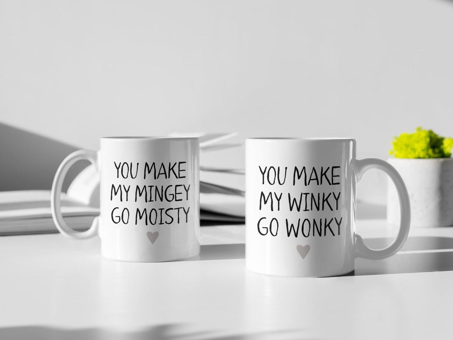 A pair of white ceramic mug featuring the funny quotes you make my mingey go moisty and you make my winky go wonky. Printed in black ink with a grey heart to the bottom centre of the design.