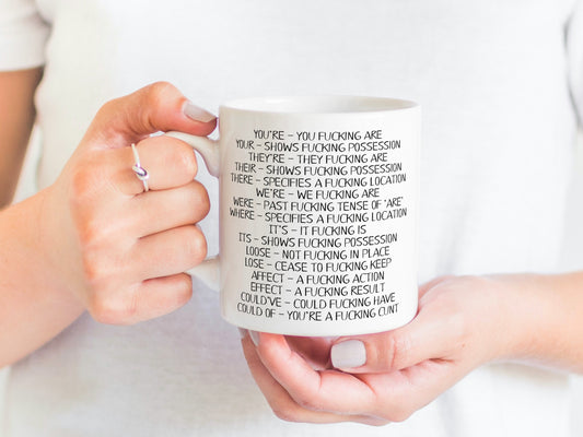 A white ceramic mug featuring funny spelling corrections such as You've - You fucking are and Your- shows fucking progression. Printed in black ink.