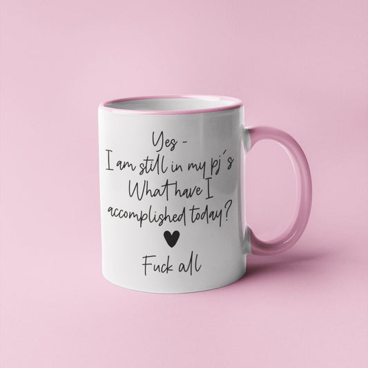 A white ceramic mug with a pink handle featuring the funny quote ‘yes i'm still in my pj's. What have i accomplished today? Fuck all’. Printed in black ink with a black heart.