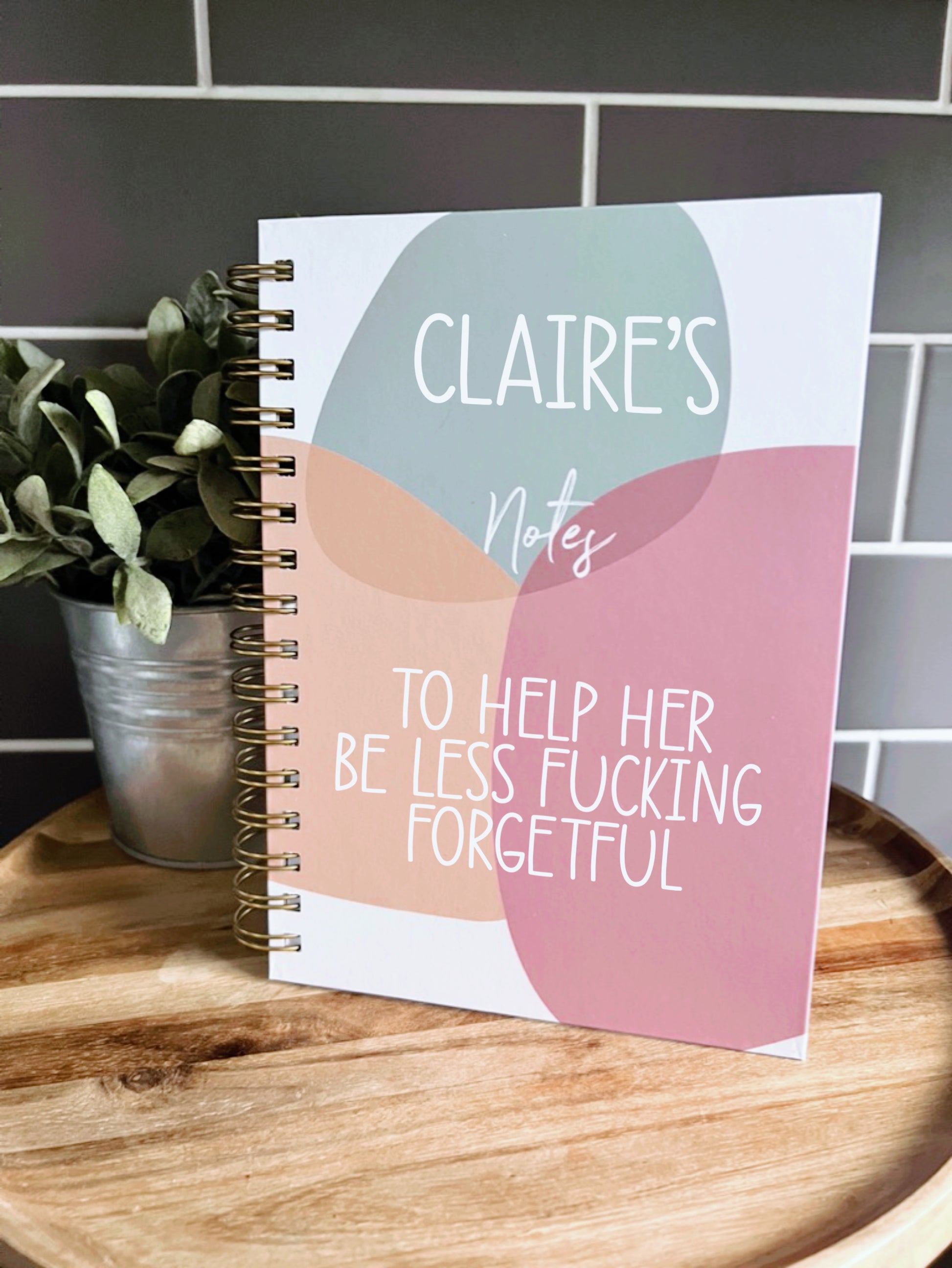 A chunky, metal binded notebook featuring the funny quote 'Claire's notes, to help her be less fucking forgetful. It has a fun circle design to the front in the colours sage, orange and pink. 