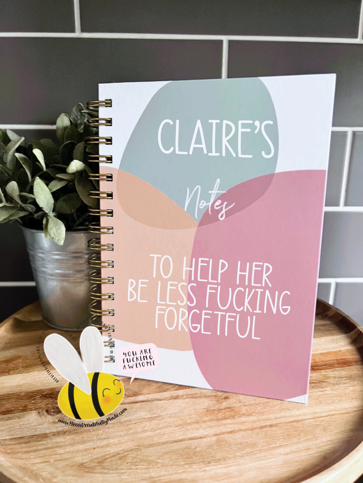 A chunky, metal binded notebook featuring the funny quote 'Claire's notes, to help her be less fucking forgetful. It has a fun circle design to the front in the colours sage, orange and pink. 