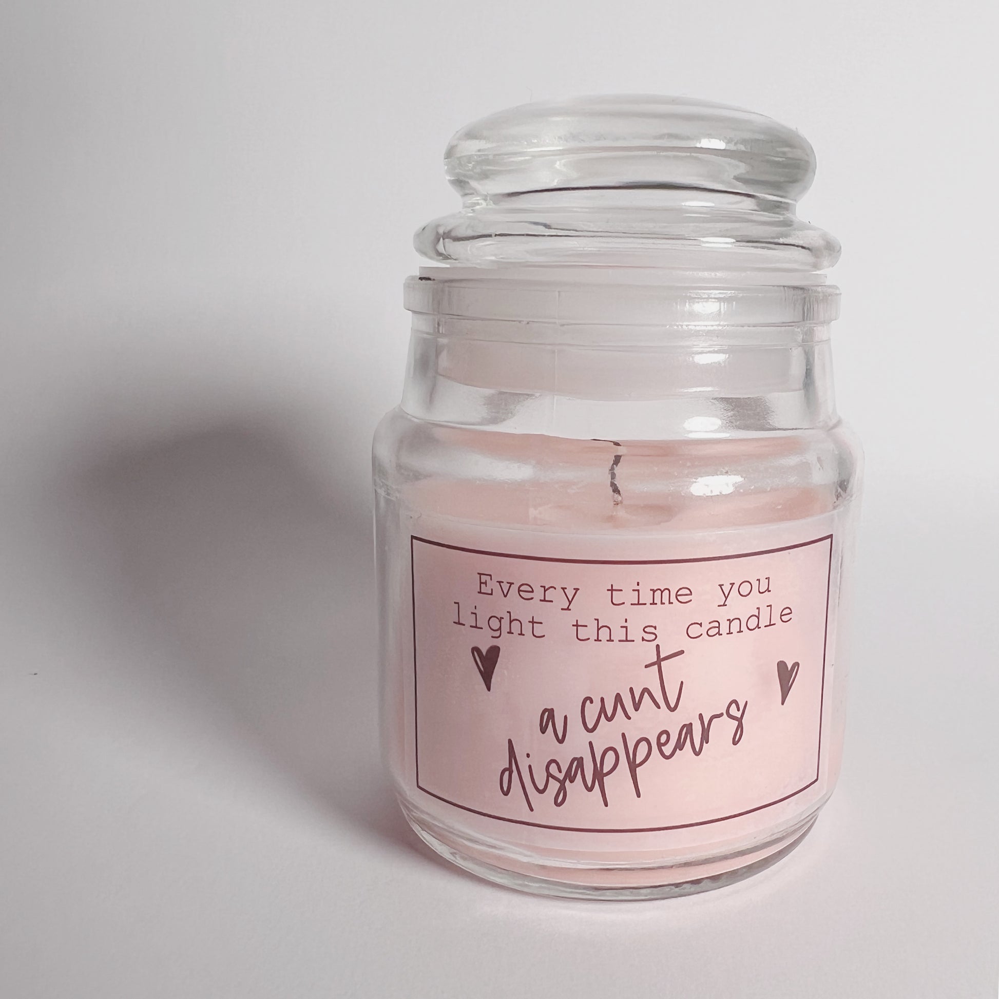 A jar candle with a lid and pink wax with a funny sticker to the front which reads, every time you light this candle a c*nt disappears.