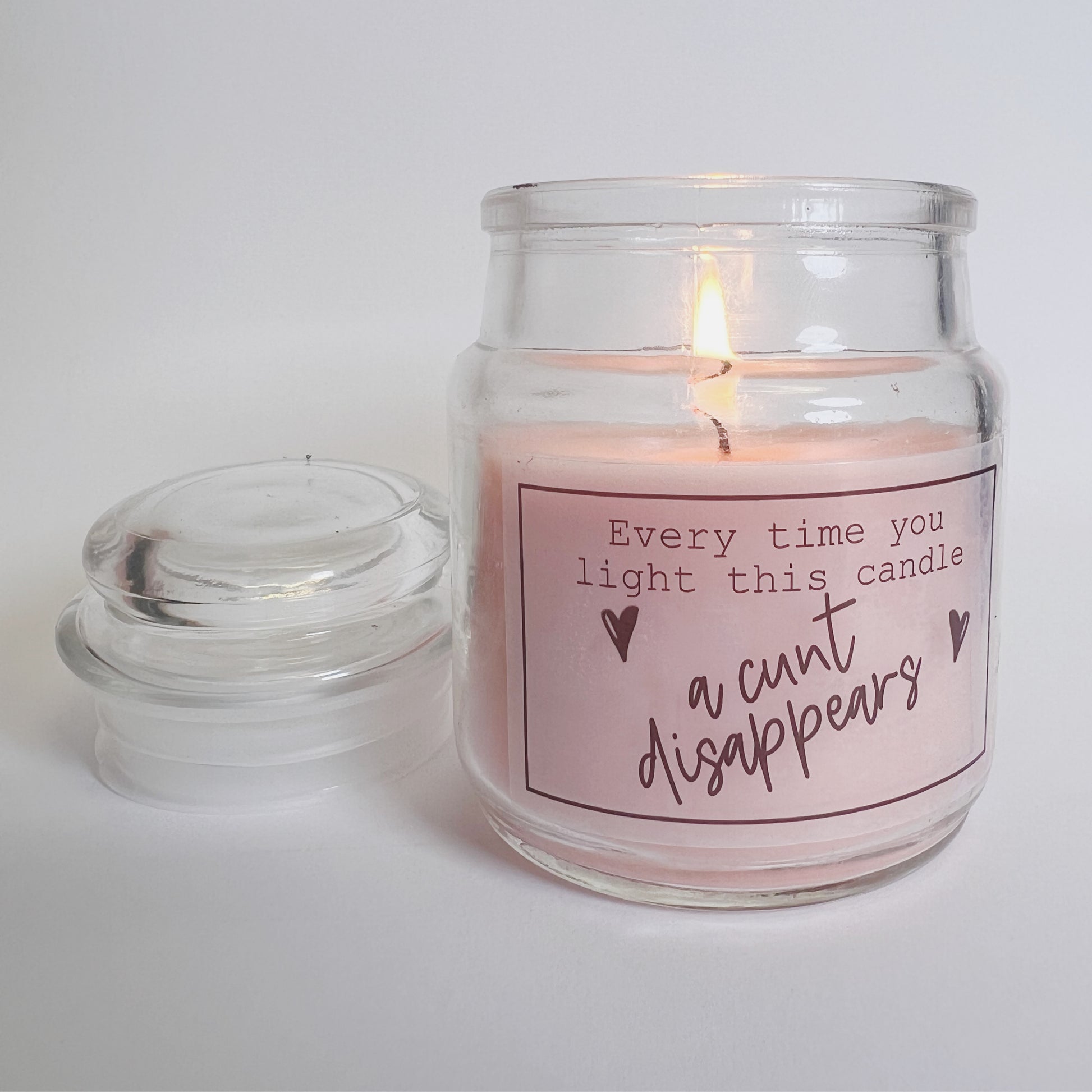 A jar candle with a lid and pink wax with a funny sticker to the front which reads, every time you light this candle a c*nt disappears.