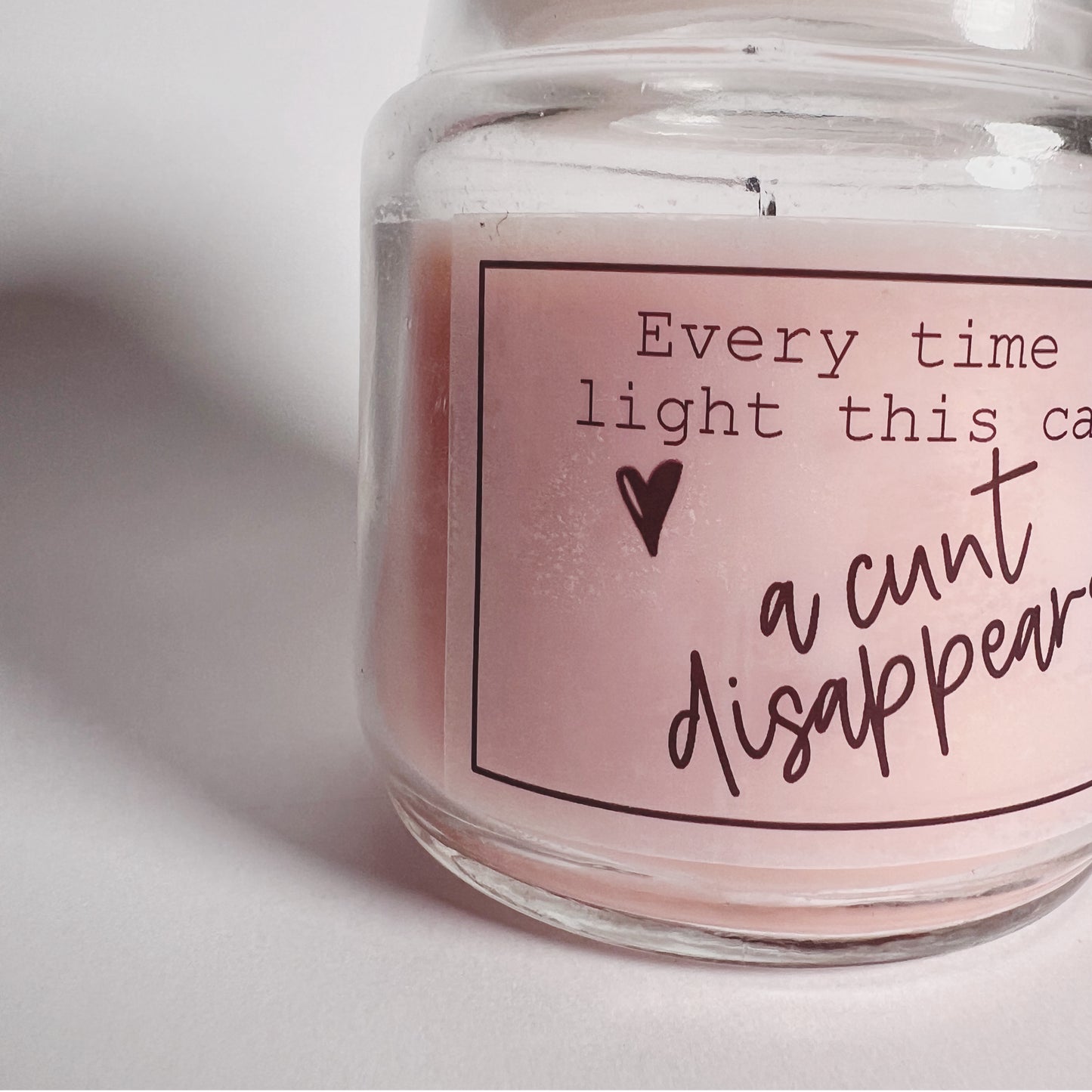 A jar candle with a lid and pink wax with a funny sticker to the front which reads, every time you light this candle a c*nt disappears.