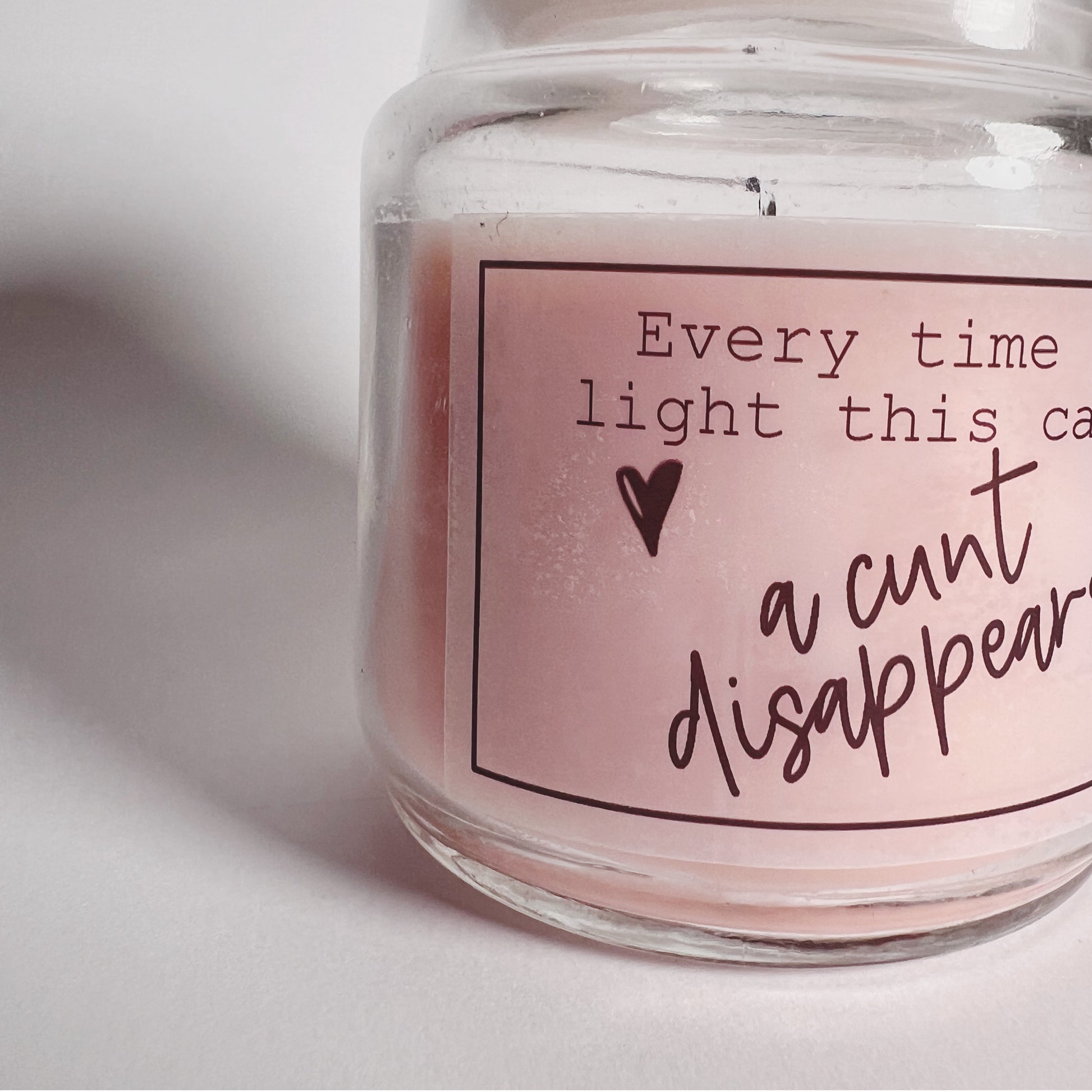 A jar candle with a lid and pink wax with a funny sticker to the front which reads, every time you light this candle a c*nt disappears.