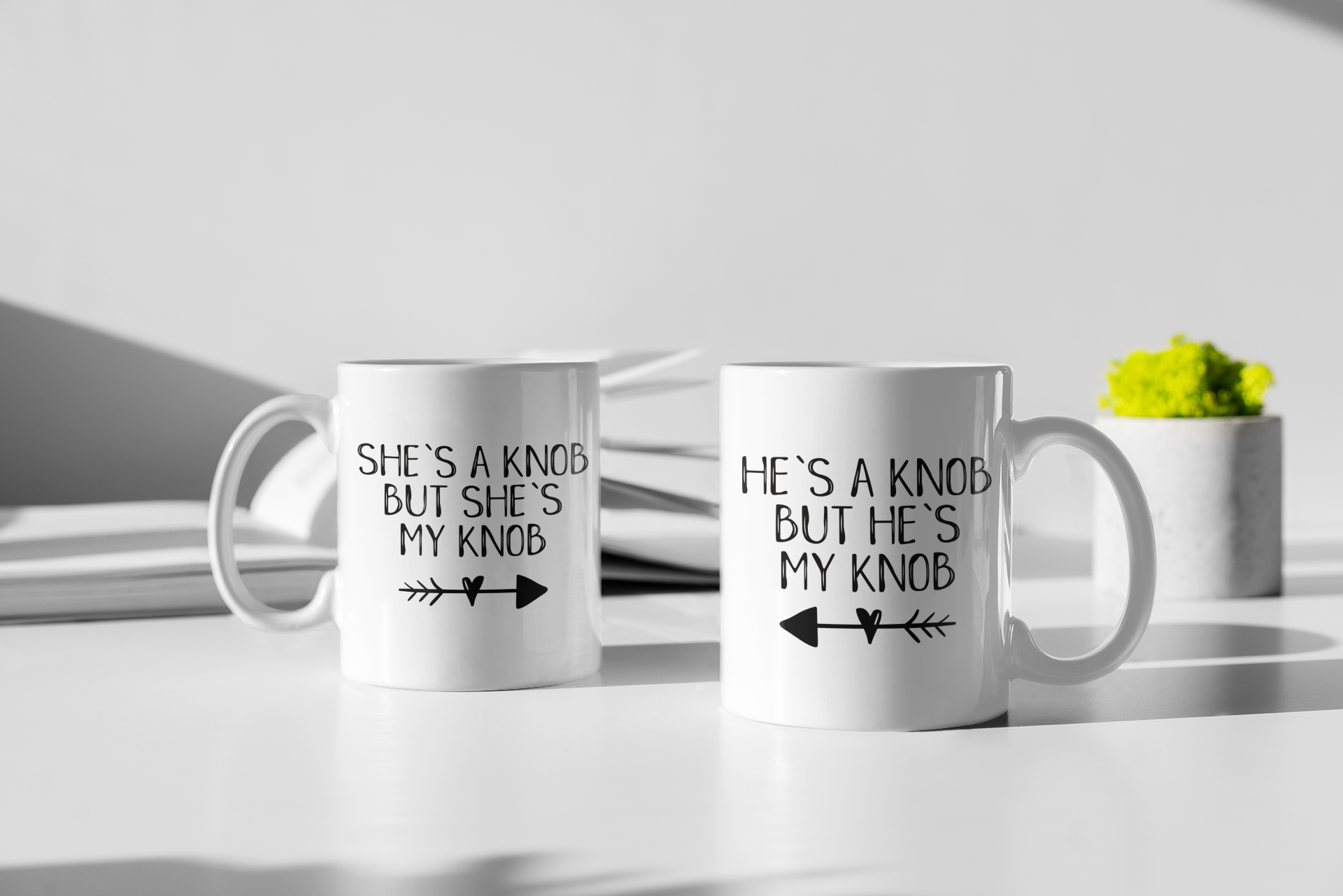 A pair of white ceramic mugs. One has 'he's a knob, but he's my knob' with an arrow underneath pointing to the left. The other has 'she's a knob, but she's my knob' with the arrow pointing to the right. Both printed to the front in black ink.