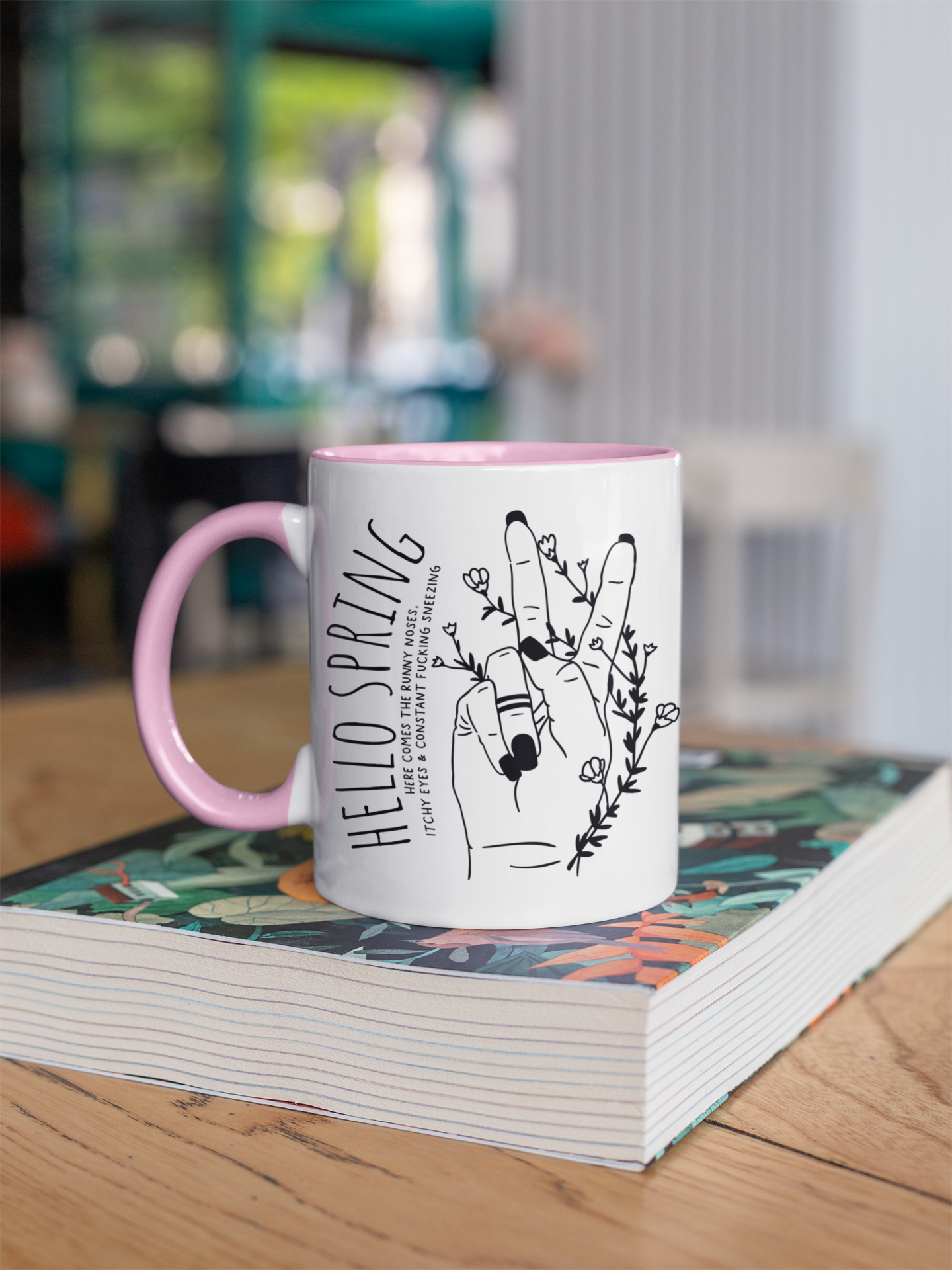 White ceramic mug with a pink handle and a cute boho style drawing of a hand sticking two fingers up, covered in twining flowers. There is a quote to the left which reads ' hello spring - here comes the runny noses, itchy eyes & constant fucking sneezing' printed in black.