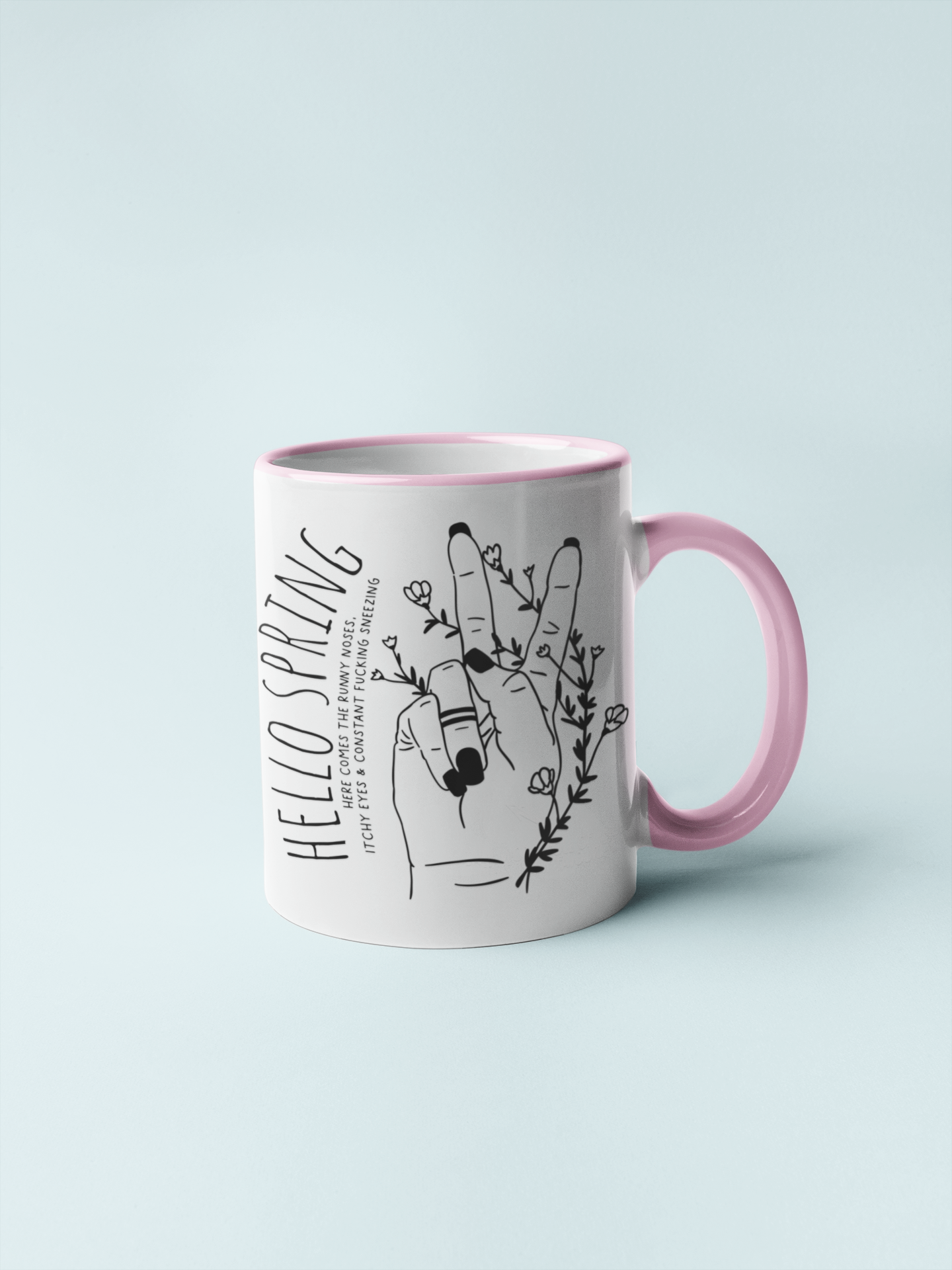 White ceramic mug with a pink handle and a cute boho style drawing of a hand sticking two fingers up, covered in twining flowers. There is a quote to the left which reads ' hello spring - here comes the runny noses, itchy eyes & constant fucking sneezing' printed in black.