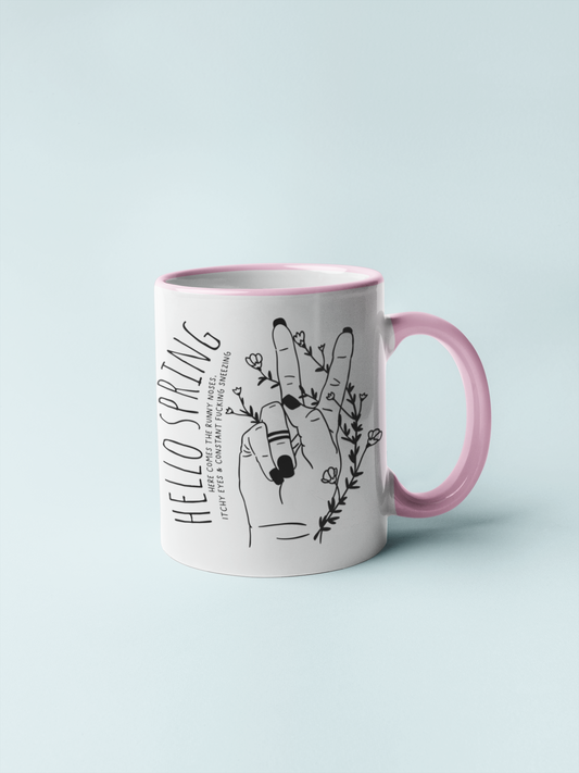 White ceramic mug with a pink handle and a cute boho style drawing of a hand sticking two fingers up, covered in twining flowers. There is a quote to the left which reads ' hello spring - here comes the runny noses, itchy eyes & constant fucking sneezing' printed in black.