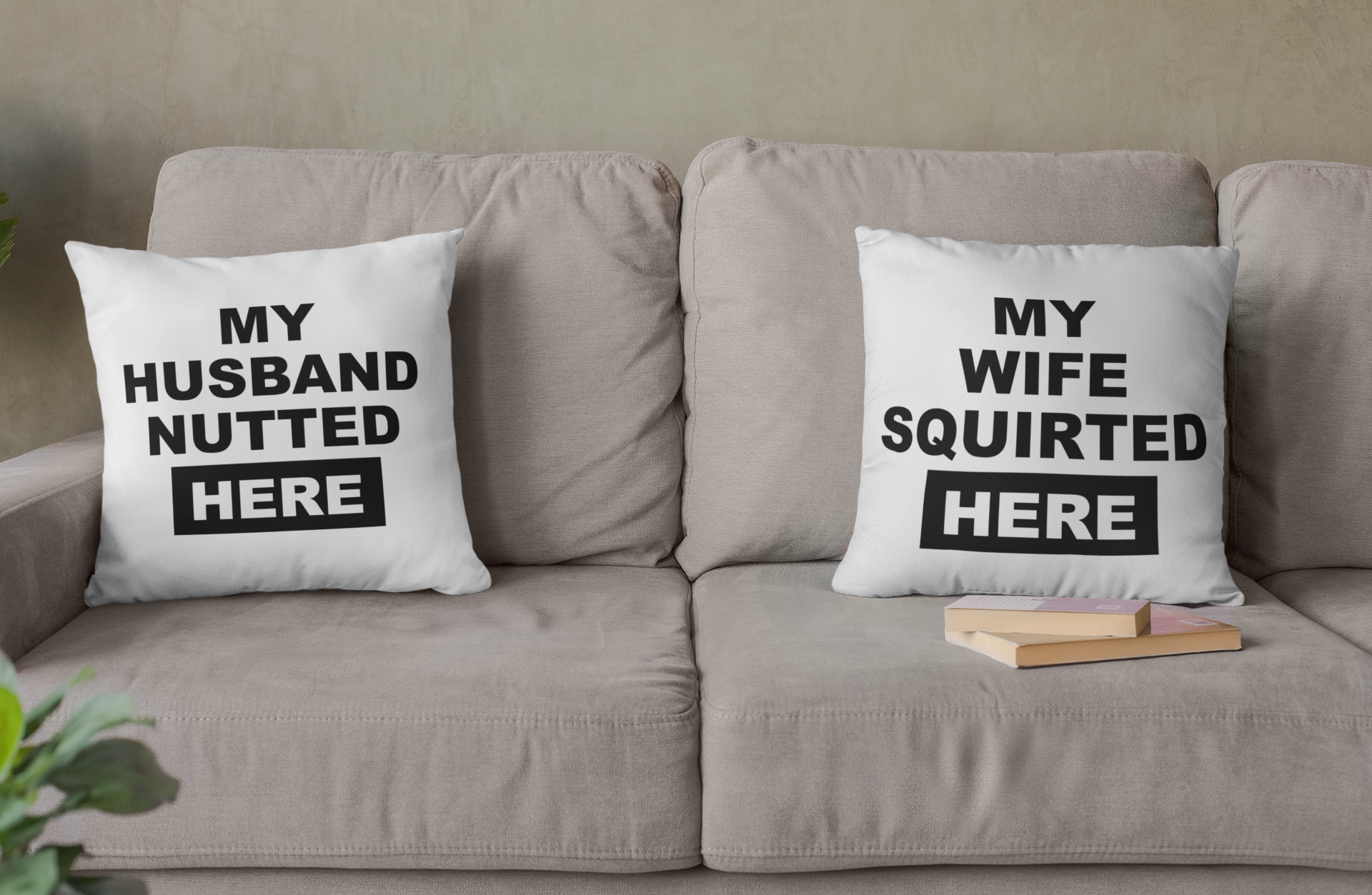 White, square cushion to the left of a sofa with bold text to the front saying 'my husband nutted here'. To the right is a maching pillow which reads 'my wife squirted here'. Both printed in black ink.