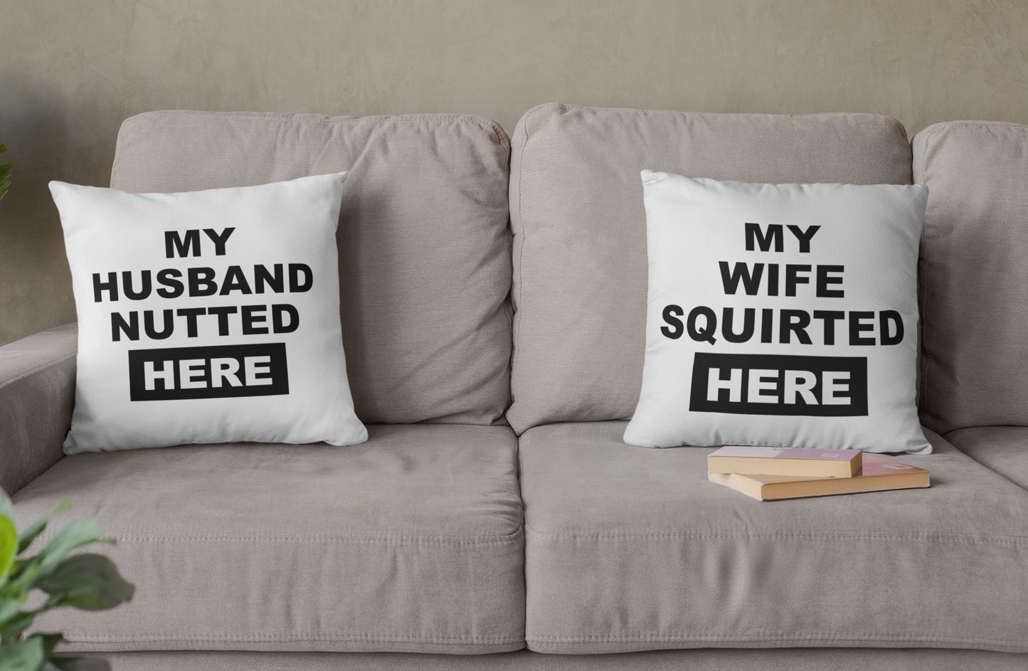 White, square cushion to the left of a sofa with bold text to the front saying 'my husband nutted here'. To the right is a matching pillow which reads 'my wife squirted here'. Both printed in black ink.