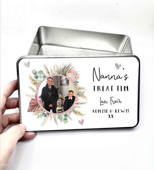 Square metal treat tin with a white slip lid. To the front of the lid it has a colourful floral design wreath with a personalised photo of 2 children in the middle. To the right of the flower is the words 'nanna's treat tin, love...'. Printed in black ink.