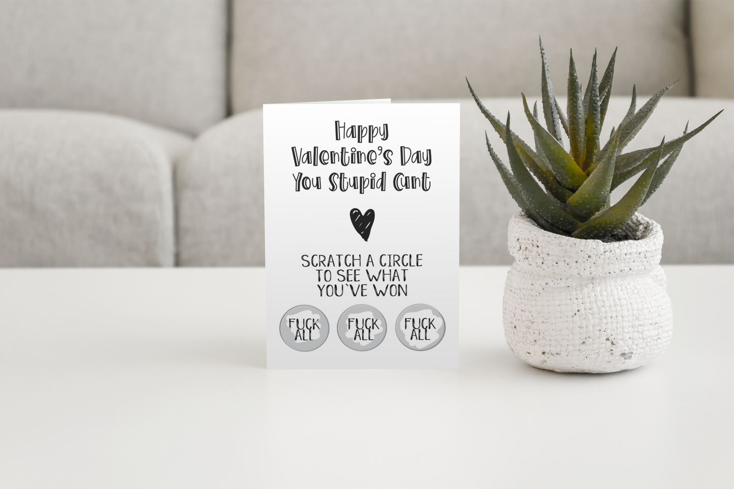 White vertical greetings card saying 'happy valentine's day you stupid c*nt' with a heart underneath. Then below is 'scratch a circle to win a prize'. To the bottom are 3 scratch stickers revealing the prizes. All printed in black ink.